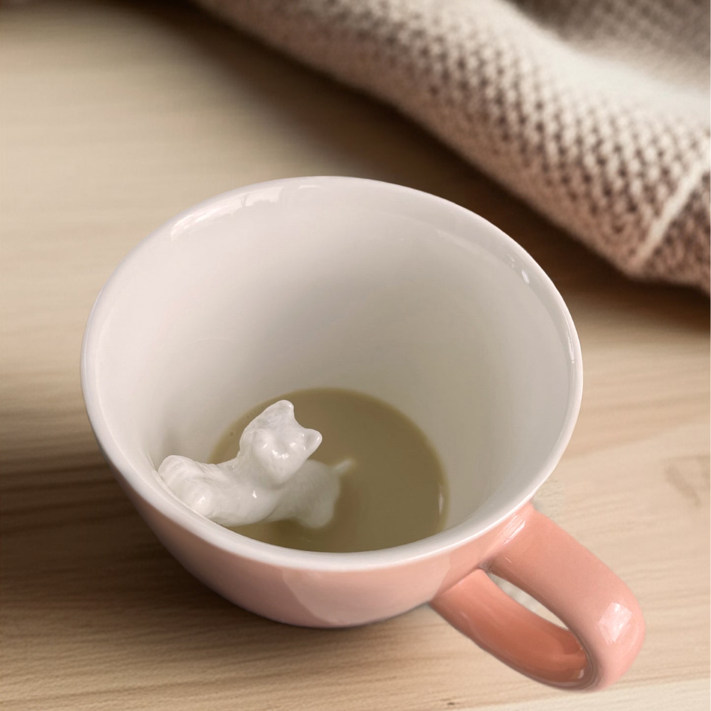 Peach colored Stretching cat Creature cup partially submerged with coffee on a wooden counter with a blanket