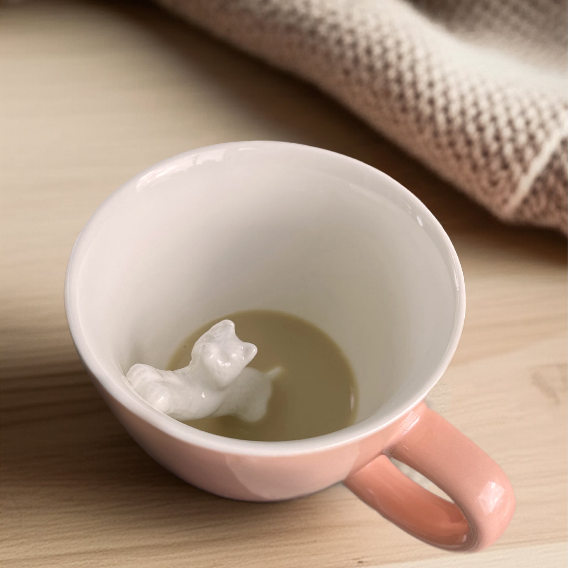 Peach colored Stretching cat Creature cup partially submerged with coffee on a wooden counter with a blanket